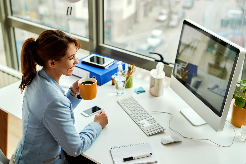 Femme d'affaire assise devant son bureau souriante et buvant un café