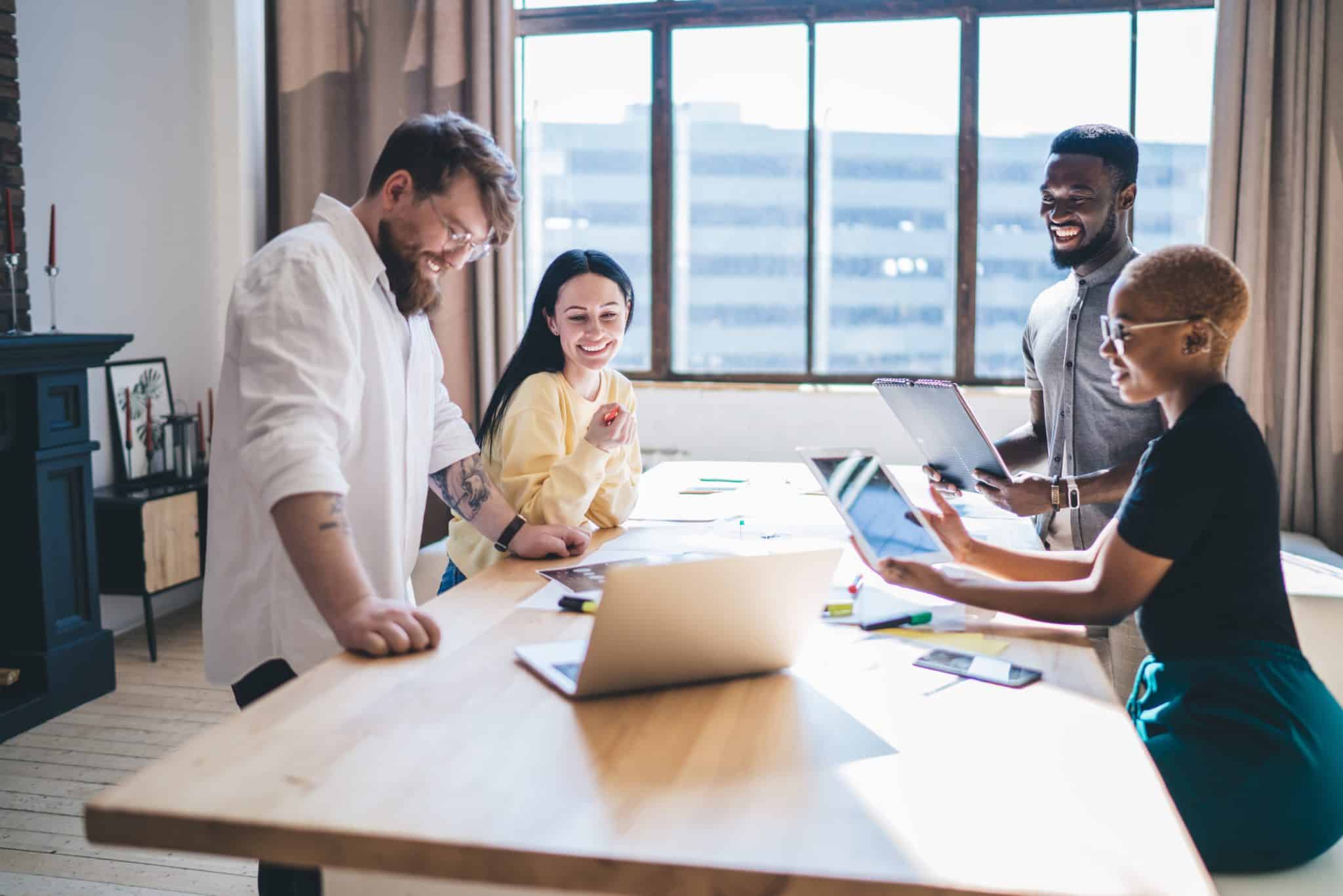 Groupe de quatre personnes souriantes travaillant autour d'une table sur des ordinateurs et tablettes dans le cadre d'un projet informatique