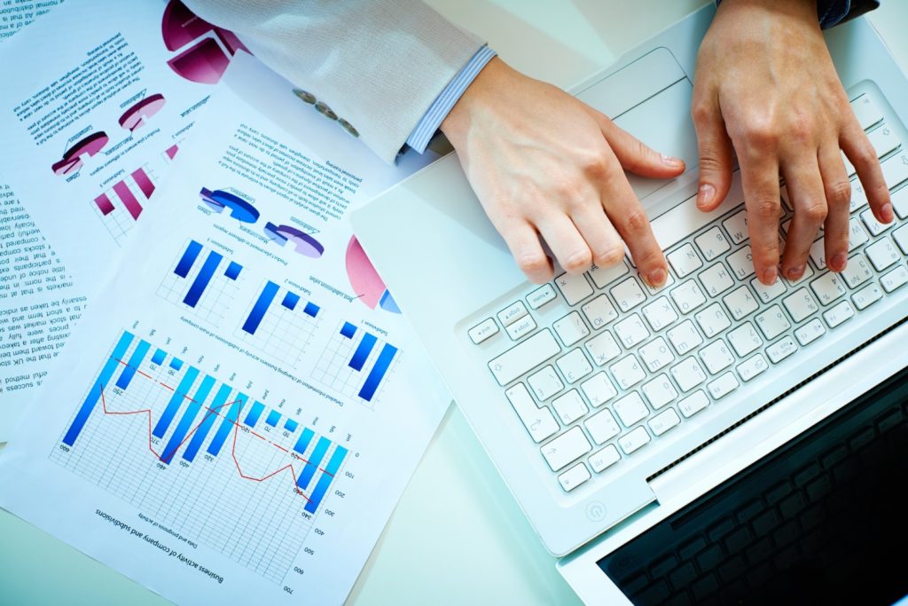 Point de vue du dessus d'un bureau avec un ordinateur portable et des fiches avec des statistiques imprimées. On voit également des mains qui écrivent sur le clavier de l'ordinateur.
